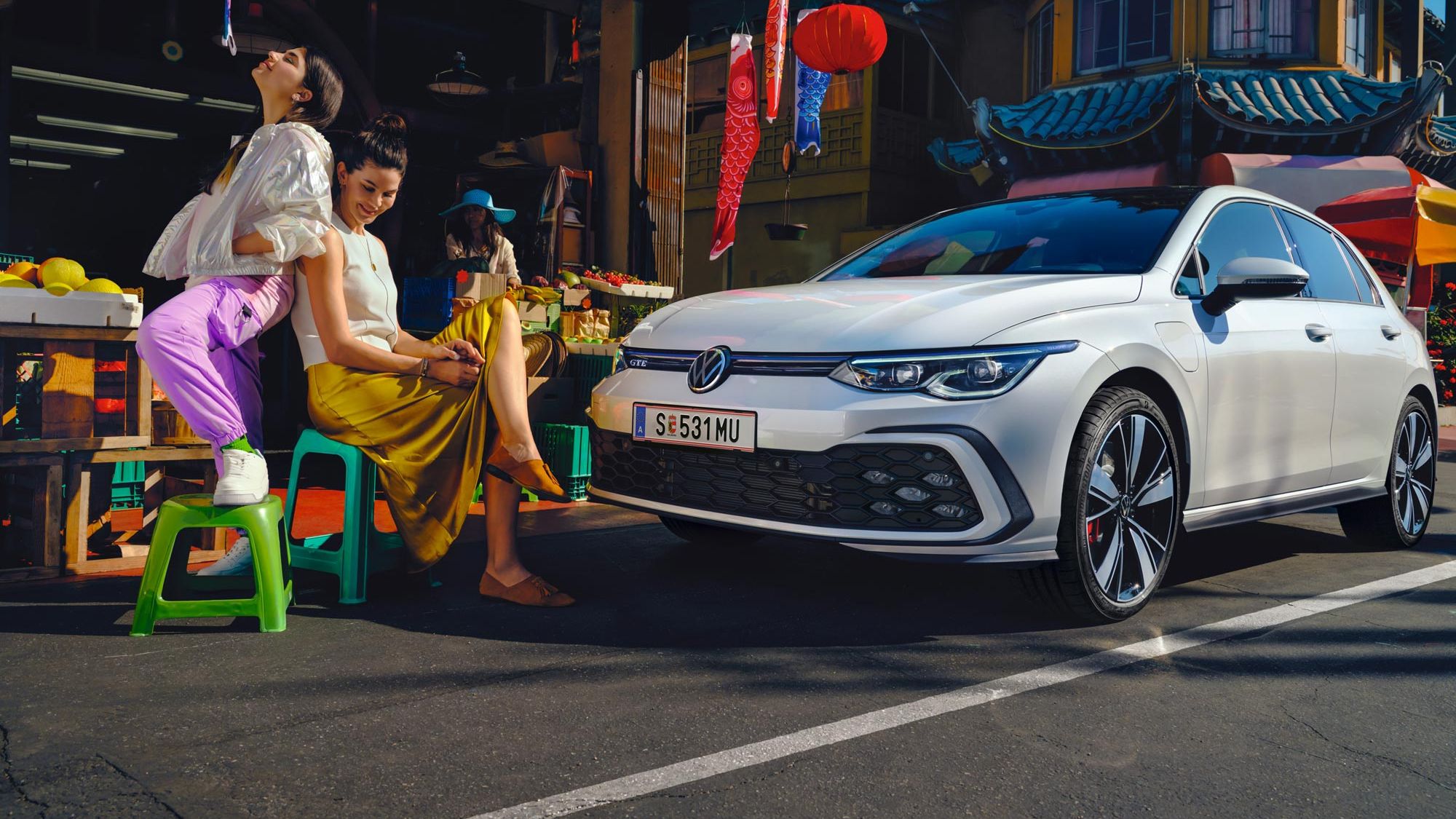 VW Golf GTE steht in Innenstadt mit asiatischen Elementen vor einem Gemüsestand, daneben sitzen zwei junge Frauen.