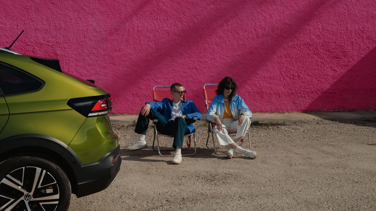 VW Taigo Heck in Grün, dahinter sitzen zwei Personen auf Campingstühlenht auf einem Parkplatz, ein Mann steht davor und blickt auf die Motorhaube.