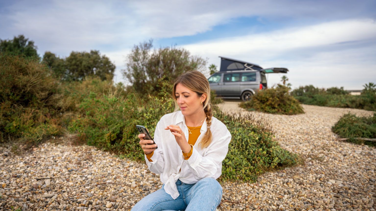 Eine Frau sitzt im Freien am Boden und blickt auf ihr Smartphone, im Hintergrund ein VW California