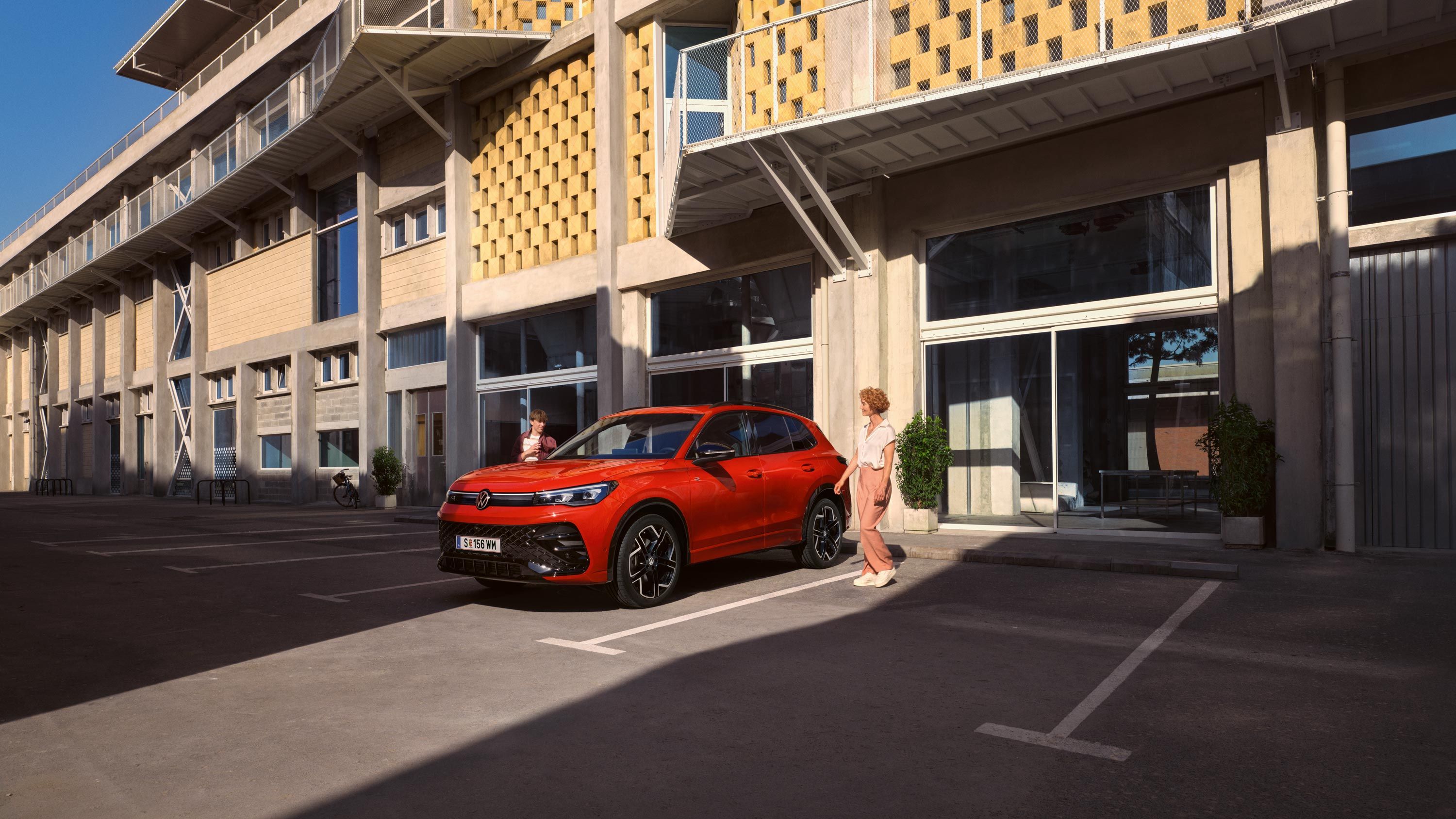 Roter VW Tiguan in Seitenansicht steht vor einem Gebäude, eine junge Frau geht auf der Fahrerseite vorbei.