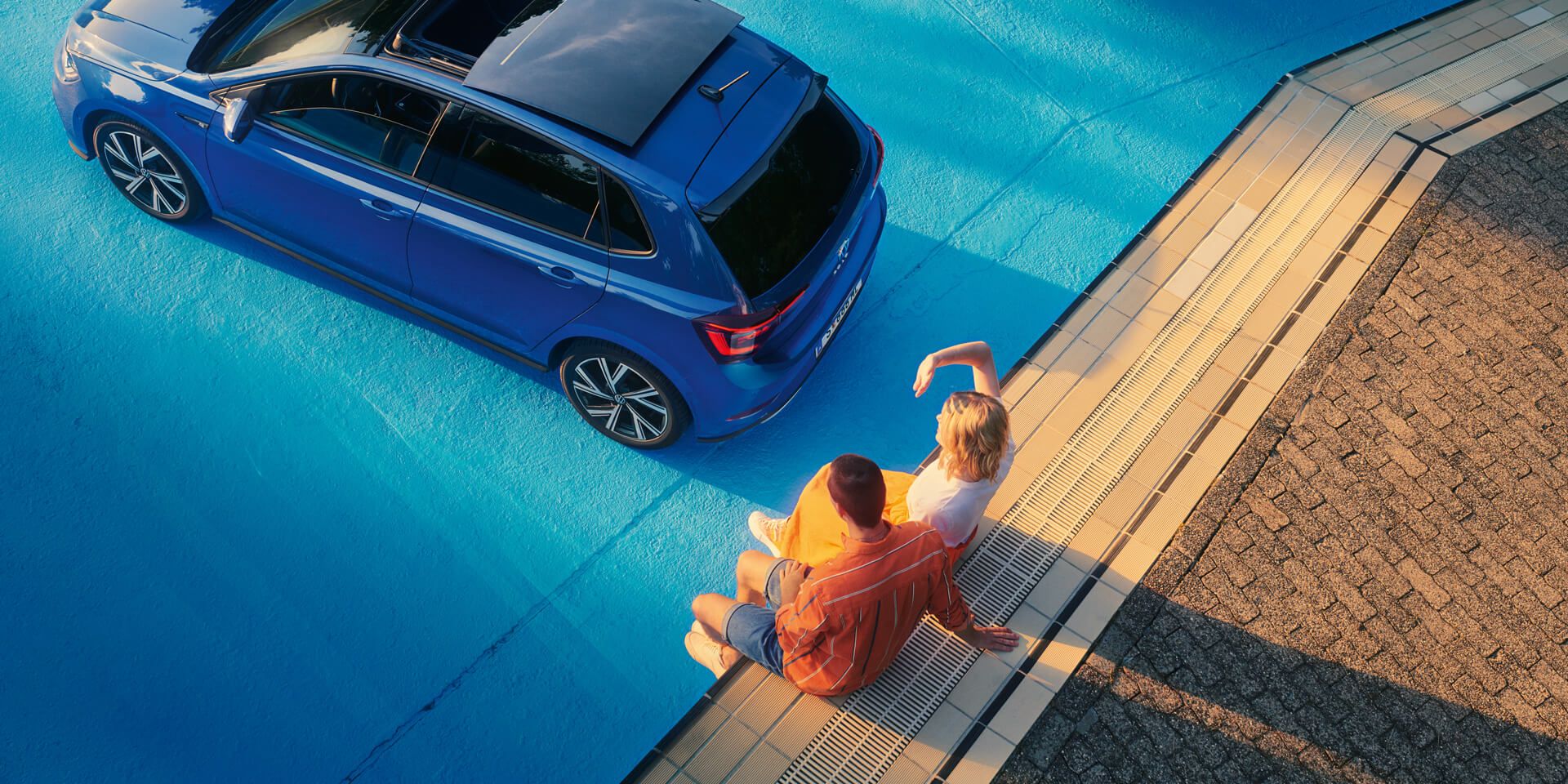 Zwei Personen sitzen am Rand eines Pools hinter einem blauen VW Polo R-line