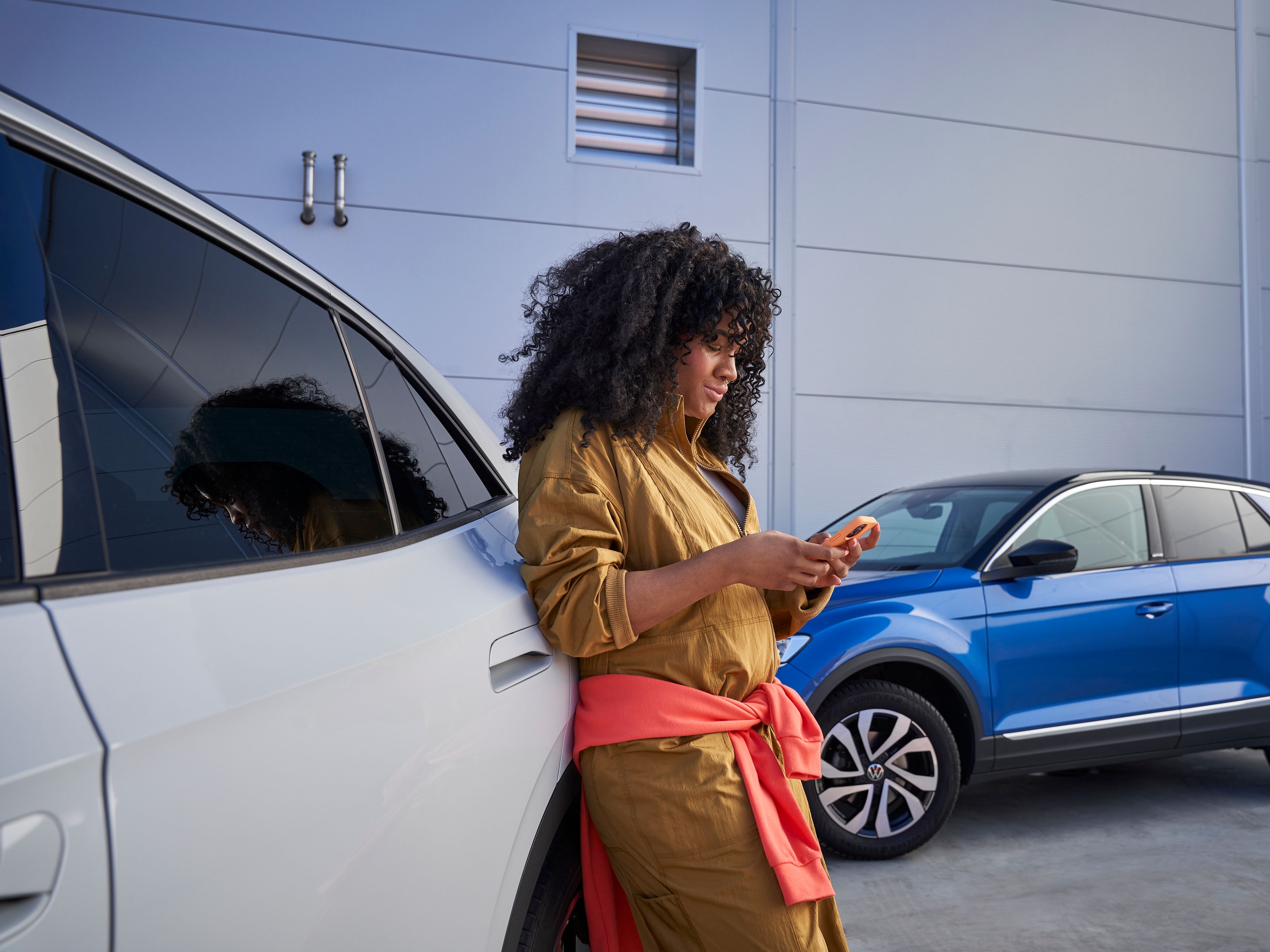 Frau lehnt an Volkswagen und blickt auf ihr Smartphone, im Hintergrund sieht man einen blauen T-Roc