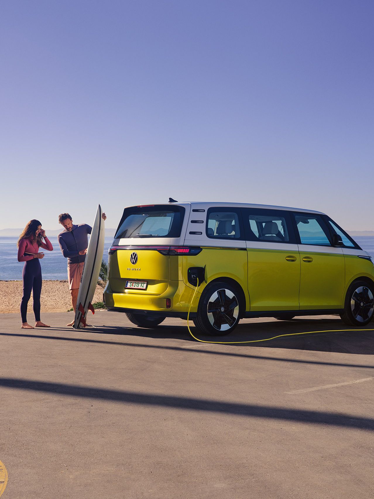 Der VW ID. Buzz parkt am Strand, davor zeigt ein Mann seiner Tochter ein Surfboard.