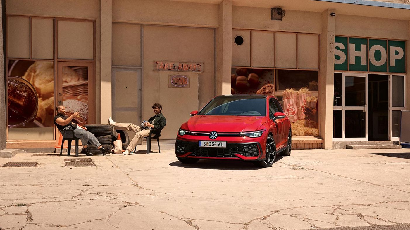 Der VW Golf GTI geparkt vor Gebäude, zwei junge Leute sitzen daneben in Stühlen