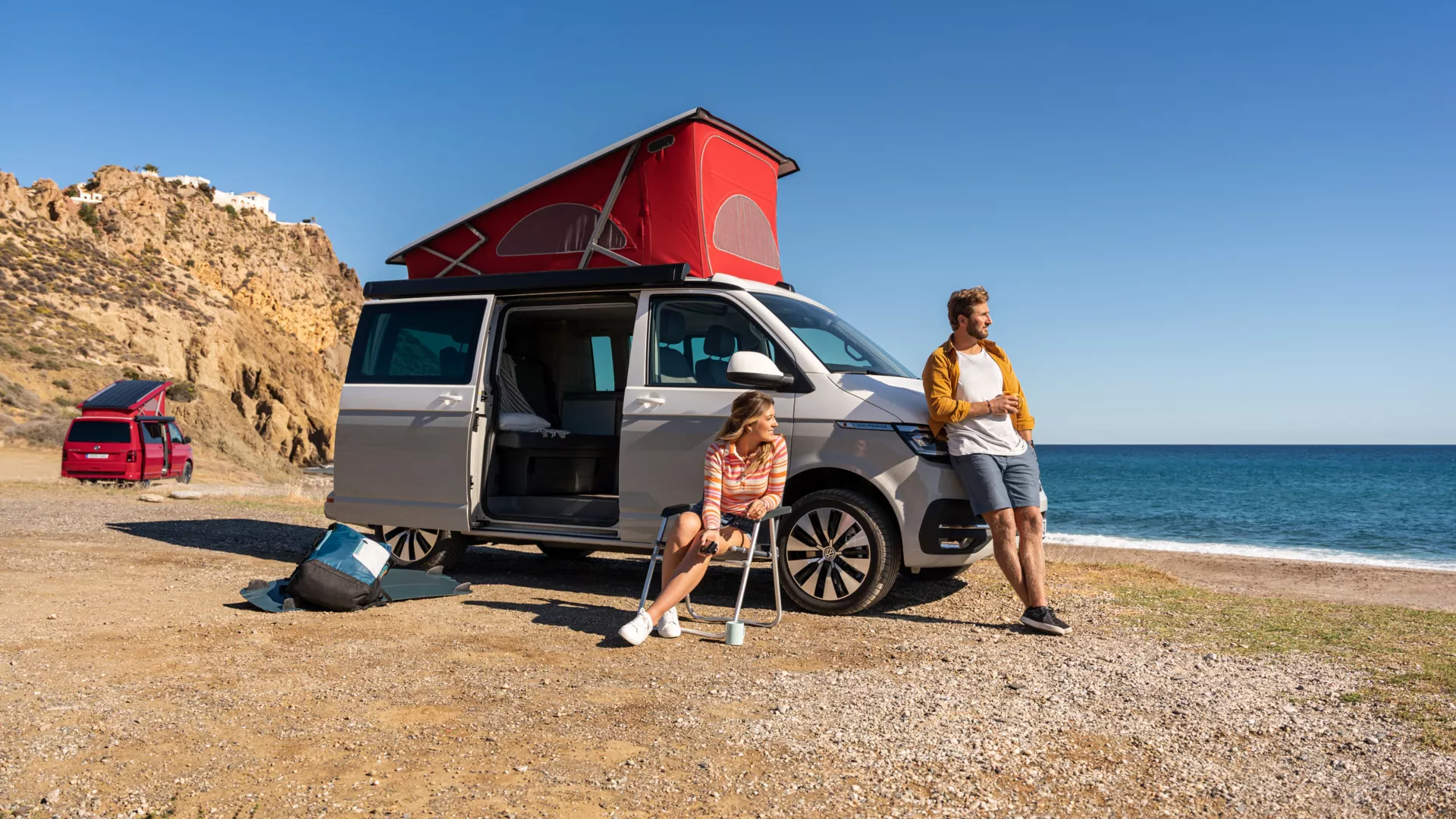 Der California 6.1 befindet sich am Strand. Im Vordergrund sind zwei Personen abgebildet, die den Camper für ihre Reise benutzen. 