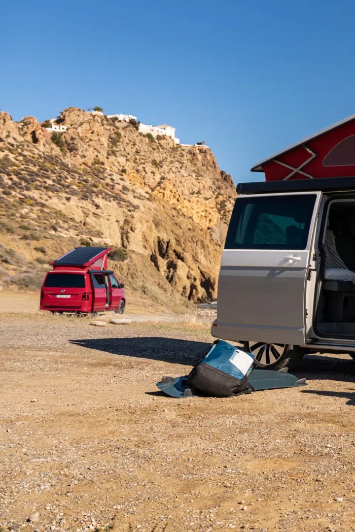Der California 6.1 befindet sich am Strand. Im Vordergrund sind zwei Personen abgebildet, die den Camper für ihre Reise benutzen. 