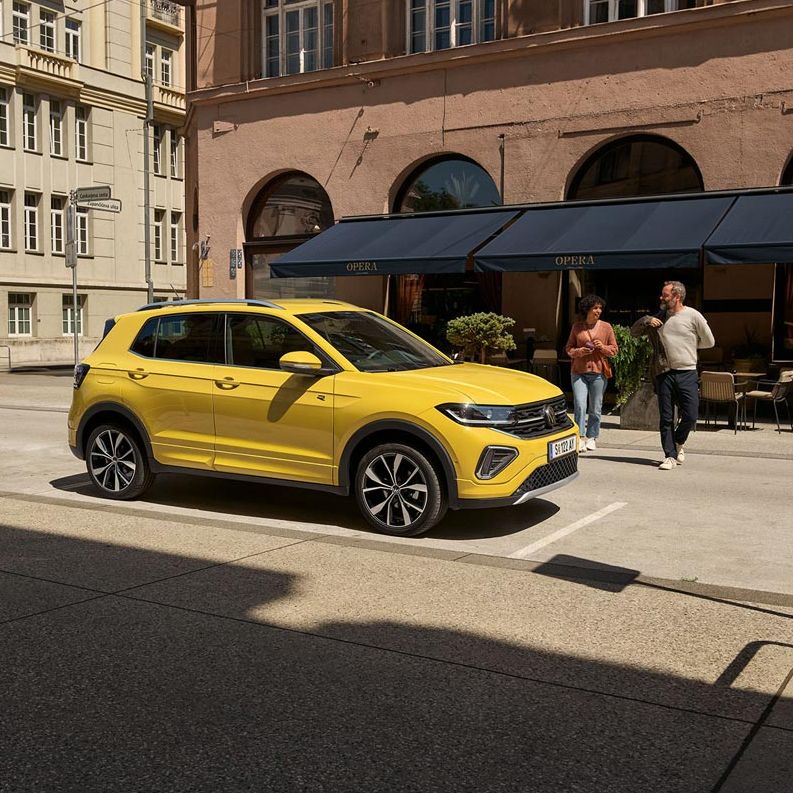 VW T-Cross in gelb parkt seitlich vor einem urbanen Straßencafé, ein Päarchen geht auf den Wagen zu.