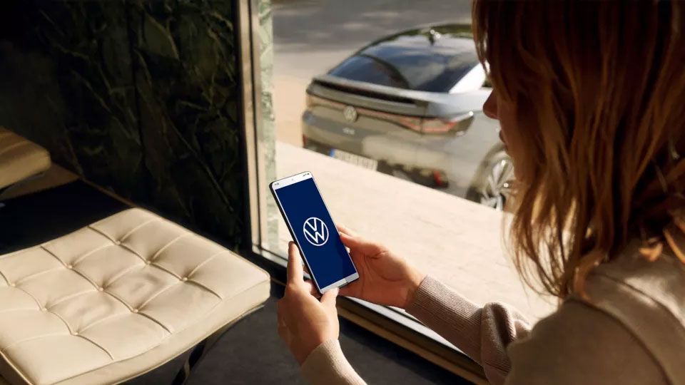 Eine Frau blickt auf ihr Smartphone, durch das Fenster ist ein VW ID. zu sehen