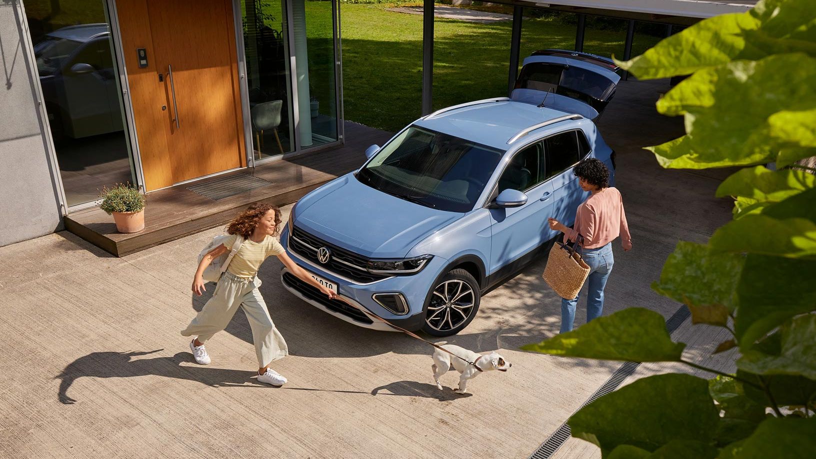 Eine Frau mit Korb und ein Mädchen mit Hund vor einem blauen VW T-Cross mit geöffnetem Kofferraum in einer Einfahrt.