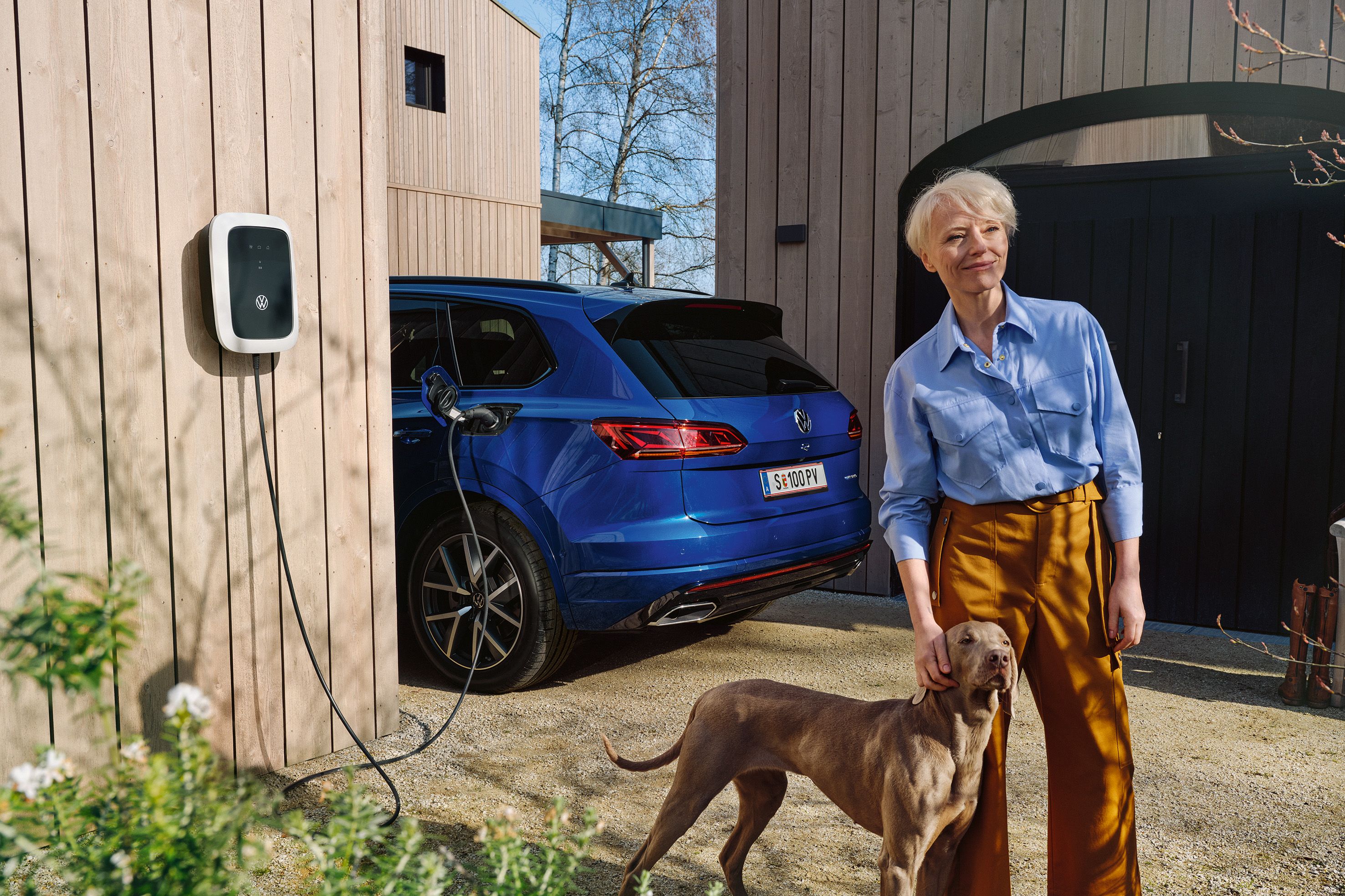 Eine Frau steht mit ihrem Hund vor einem ladenden VW Touareg R eHybrid.