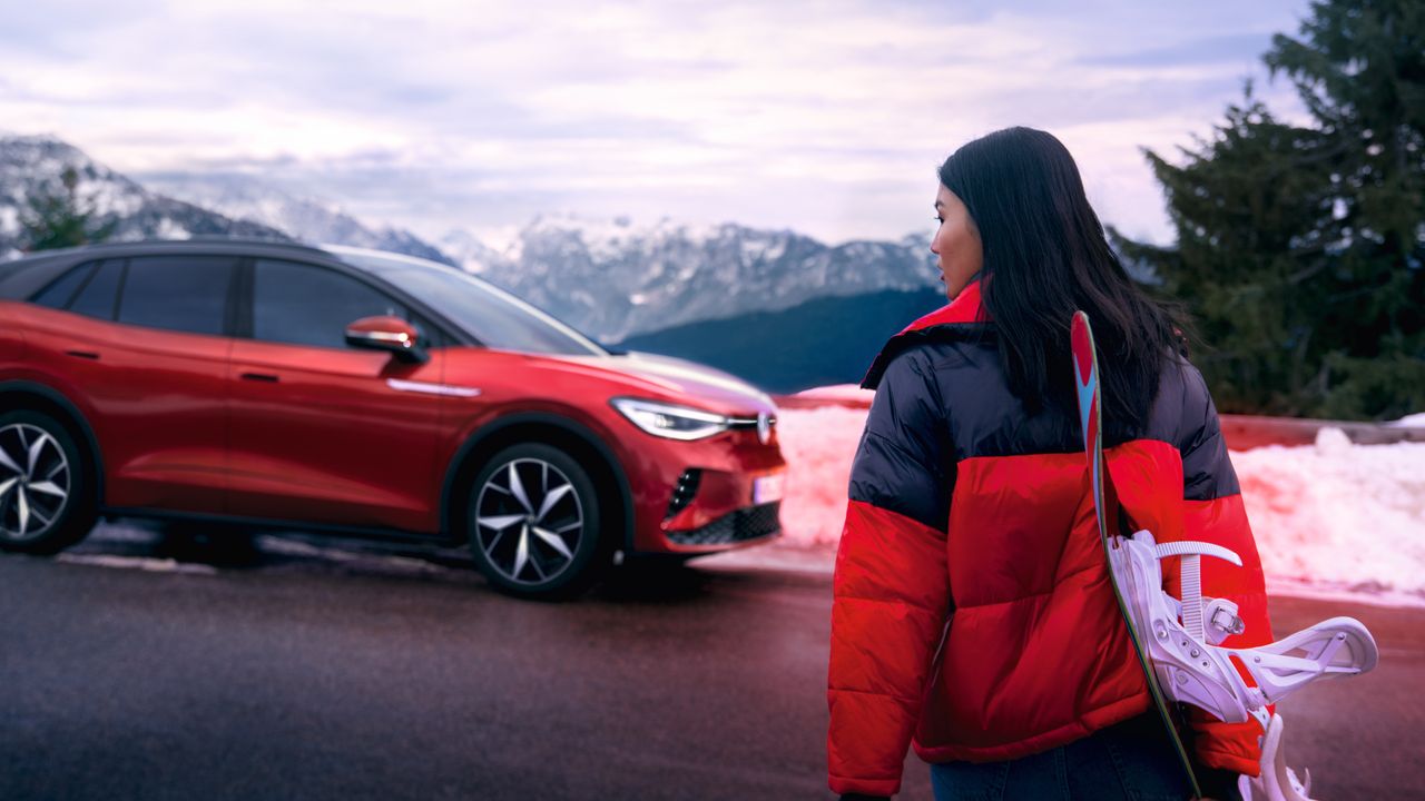 Seitenansicht eines roten VW ID.4 GTX vor Bergkulisse, im Vordergrund blickt eine Frau zum Auto