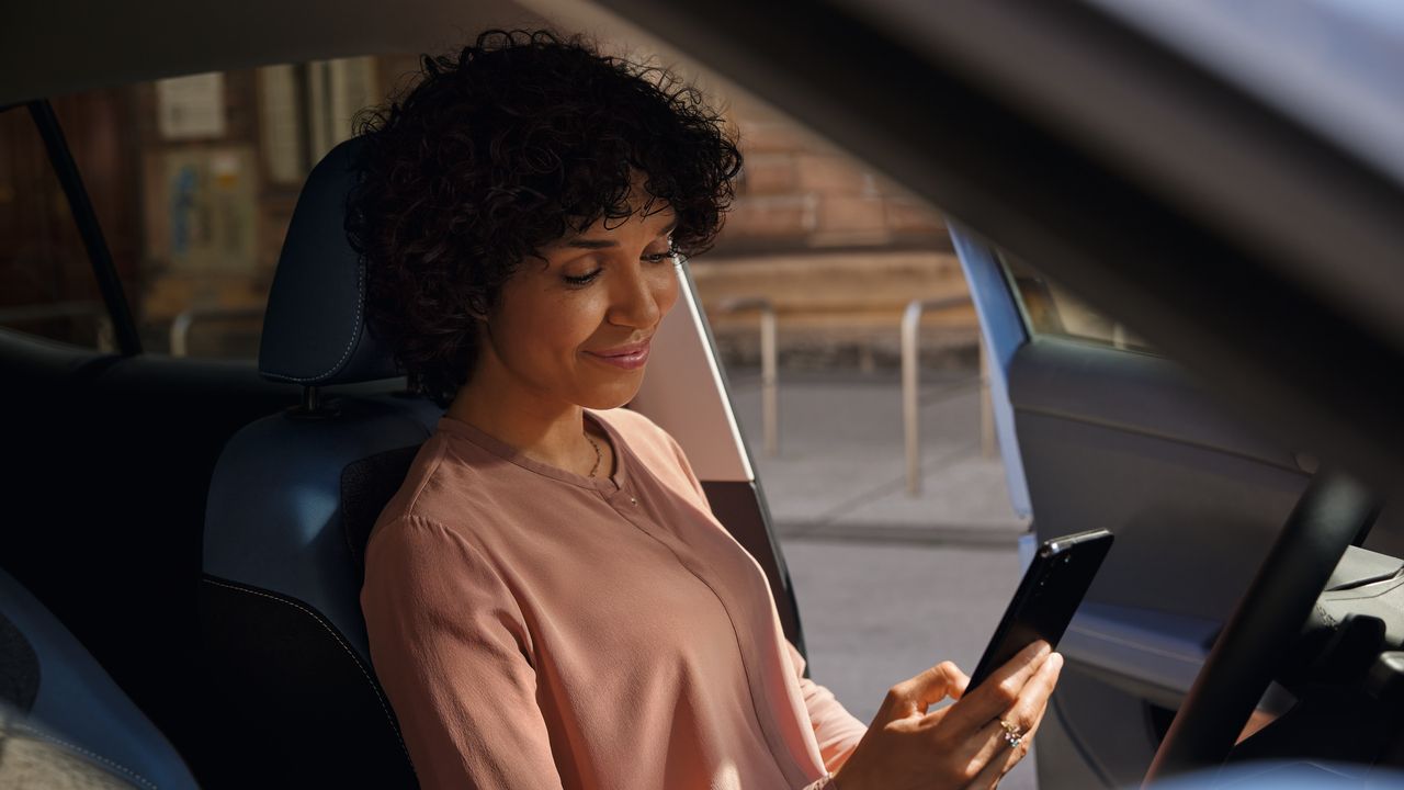 Eine lächelnde Frau schaut auf dem Fahrersitz eines geparkten VW T-Cross aufs Handy.