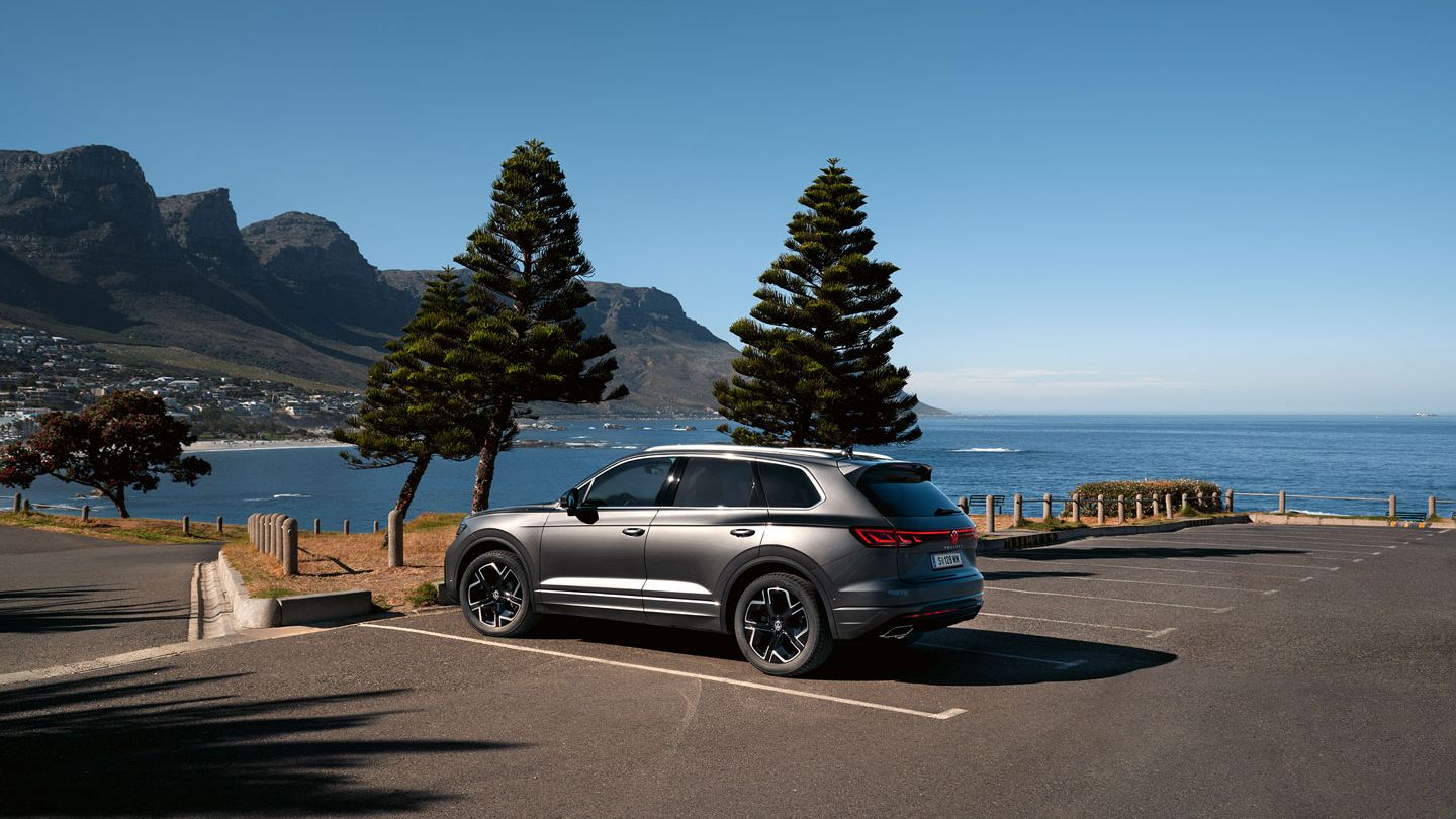Ein VW Touareg Elegance parkt in der Sonne, Ansicht von der Seite, dahinter sind das Meer und die Küste zu sehen.