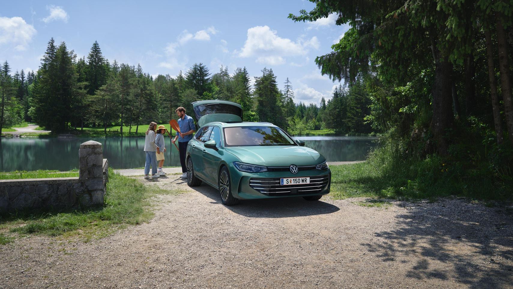 Der neue VW Passat Elegance in Maripositgrün Metallic parkt an einem Badesee mit geöffnetem Kofferraum. Ein Mann verteilt Freitzeitspielzeug an zwei Kinder.