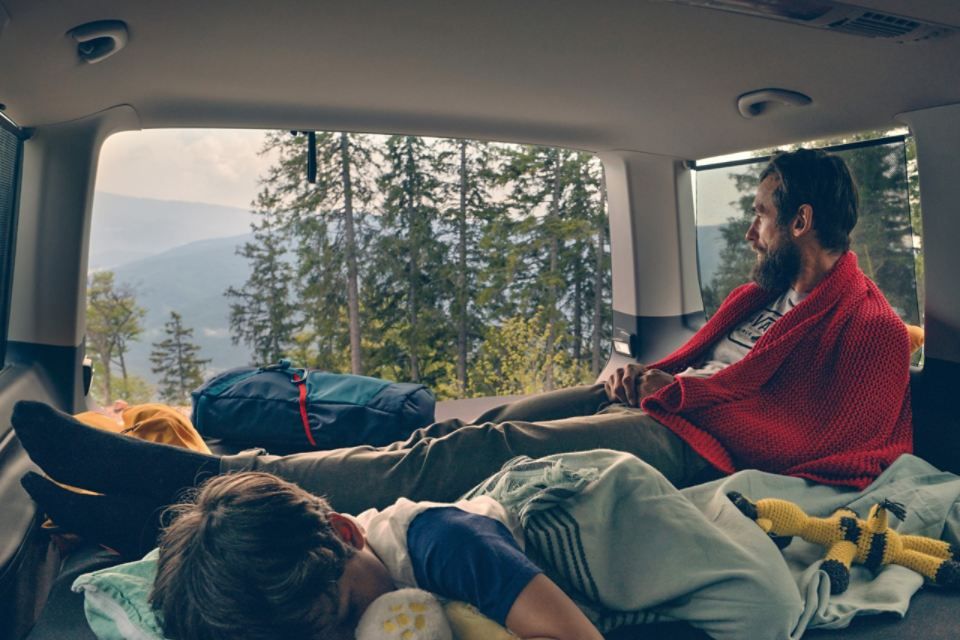 Kind schläft im VW Multivan 6.1. Vater liegt daneben und blickt aus offenem Kofferraum in Richtung Wald.