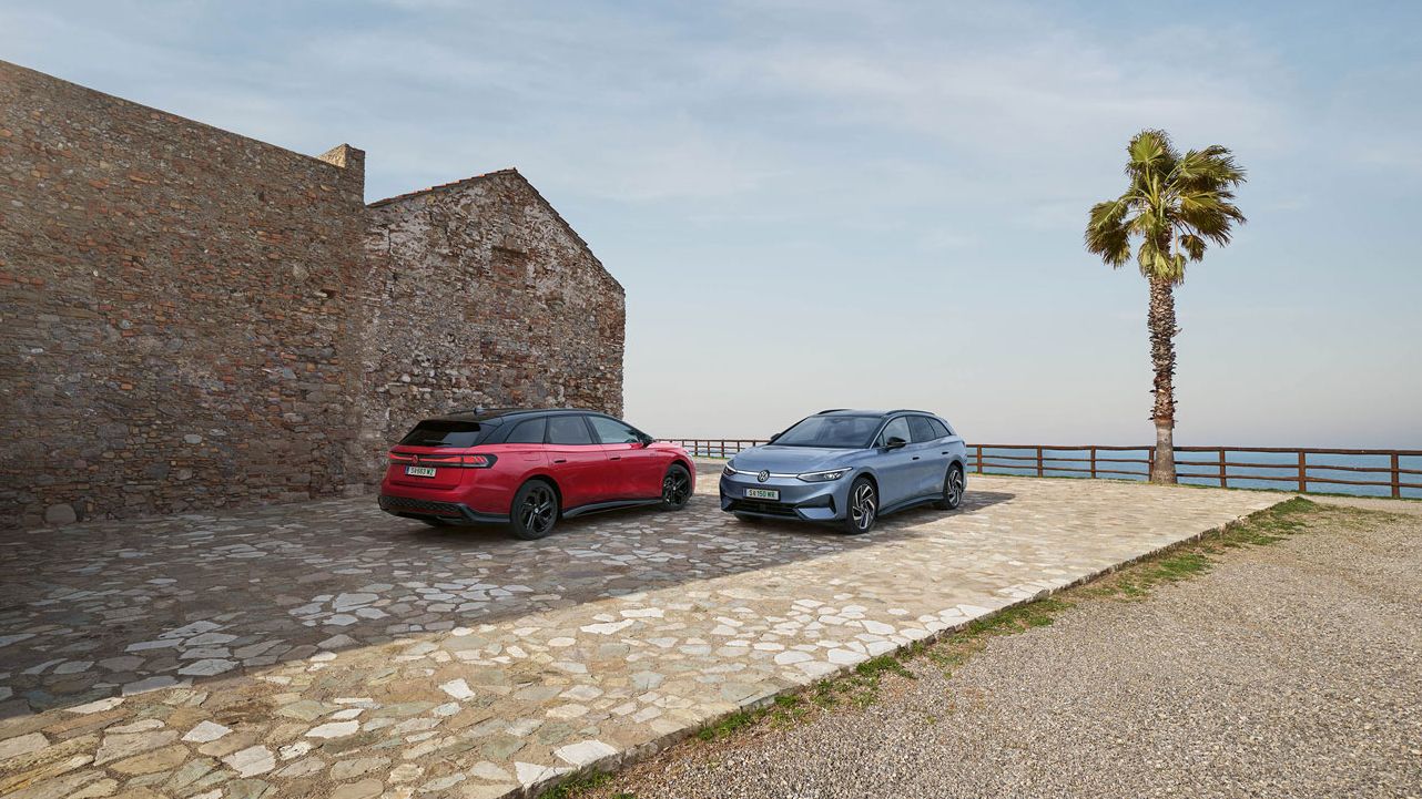 Ein VW ID.7 Tourer in Stonewashed Blue Metallic und ein VW ID.7 Tourer GTX in Kings Red Metallic geparkt an Küste neben eines Steinhaus'