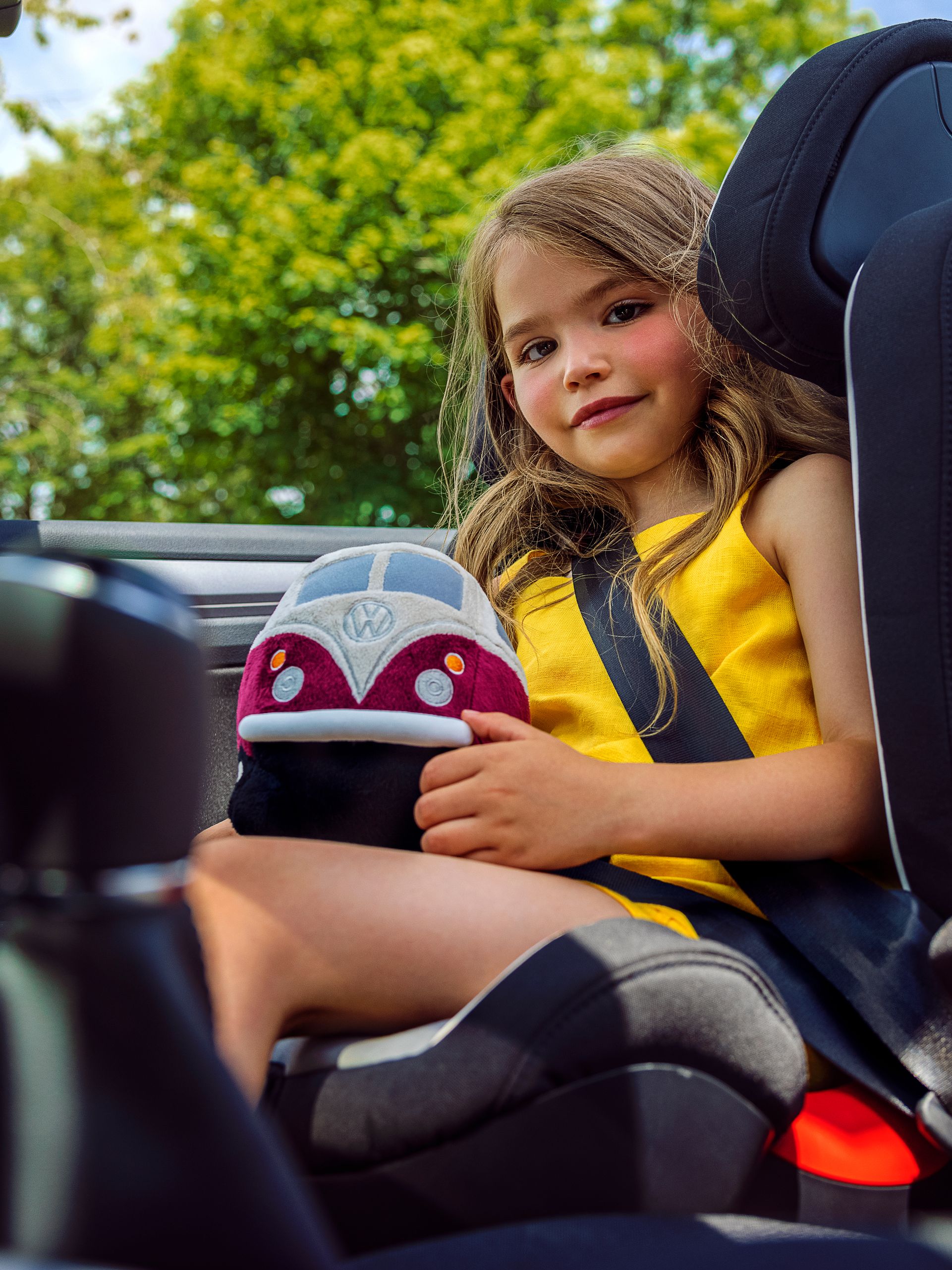 Ein Mädchen sitzt in einem VW Kindersitz und hat ein VW Bulli Kuscheltier iin der Hand