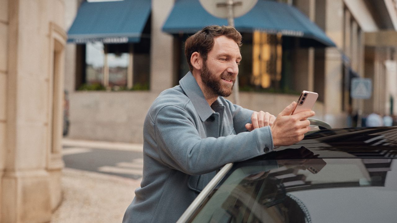 Mann lehnt an einem Volkswagen und schaut auf sein Mobiltelefon