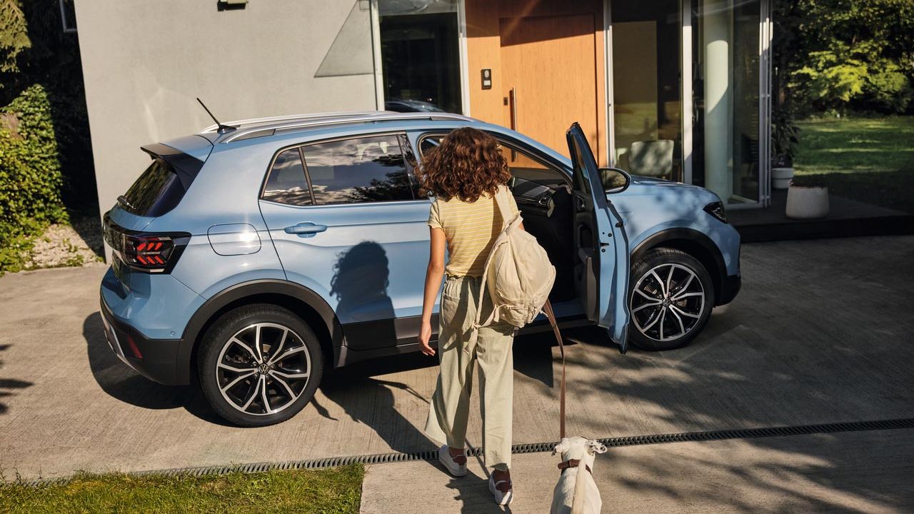 Ein Mädchen mit Hund geht auf einen blauen VW T-Cross in einer Einfahrt zu. Die Beifahrertür ist geöffnet.