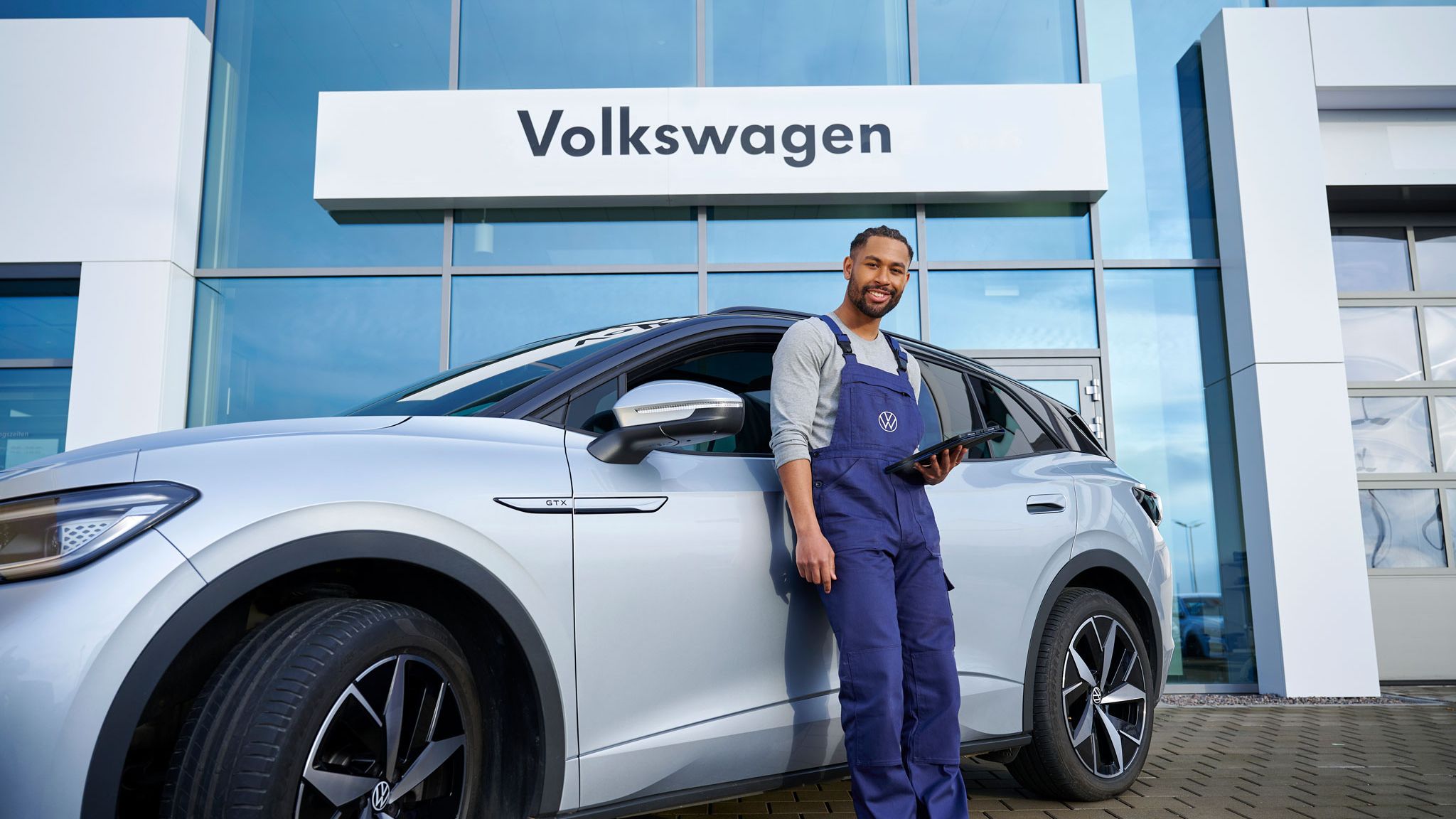 Ein Servicemechaniker lehnt an einem Volkswagen ID.4 GTX mit einem Tablet in der Hand vor einem Autohaus.