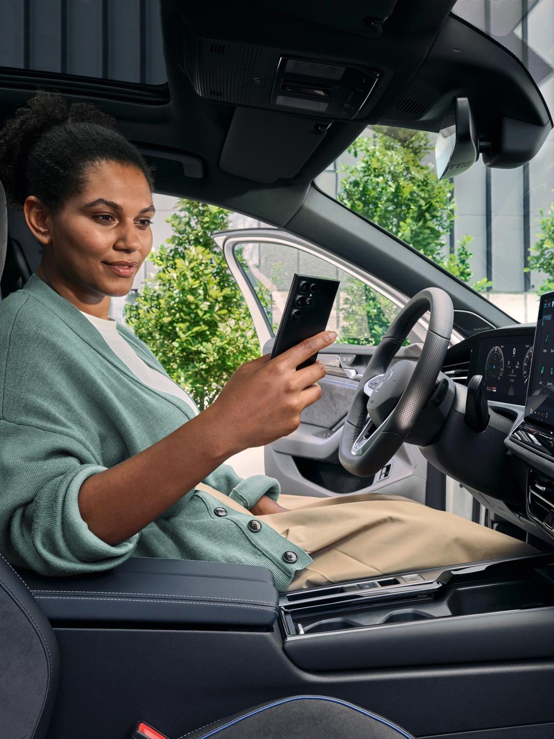 Eine Frau sitzt bei geöffneter Tür auf dem Fahrersitz des geparkten VW Passat und blickt auf ihr Smartphone.