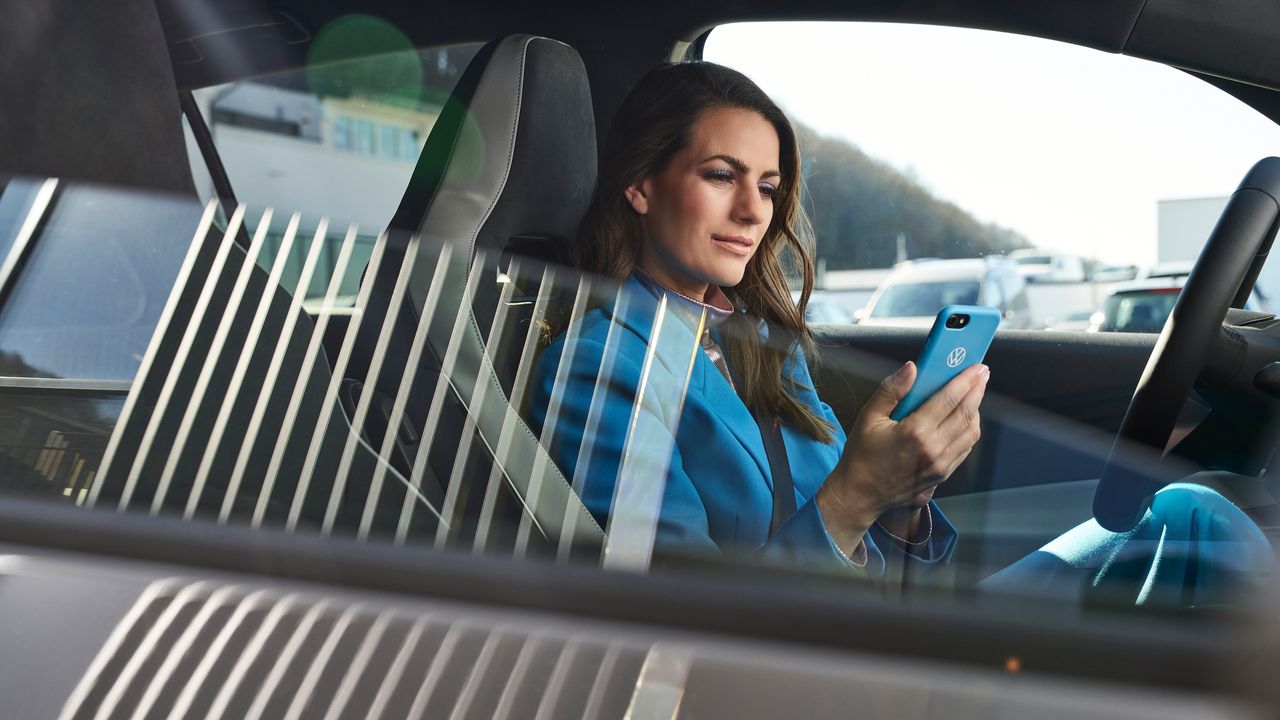 Eine in blau gekleidete Frau sitzt in einem geparkten Volkswagen mit einem Smartphone in ihrer Hand.