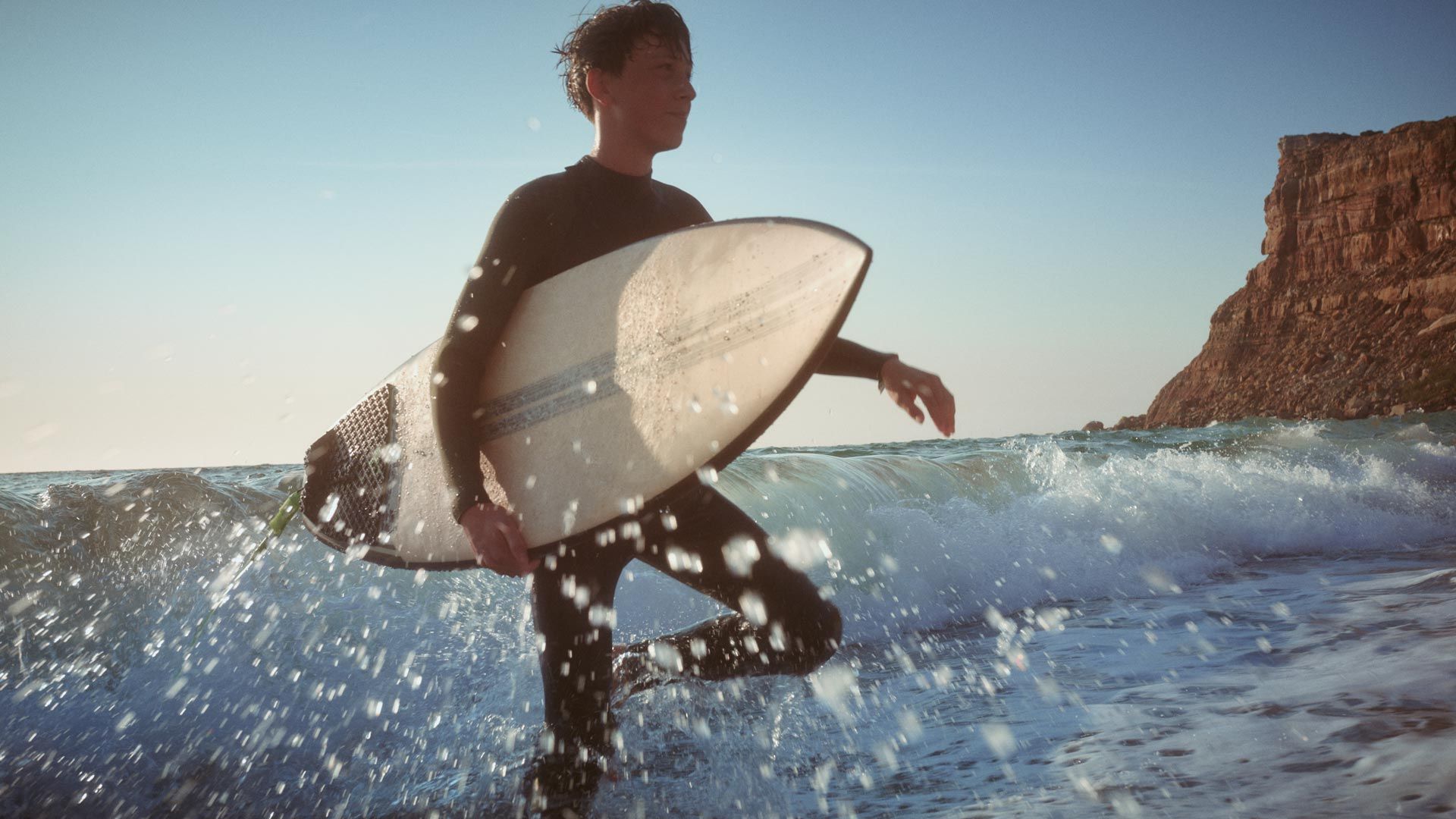 Strandszene - Ein junger Mann watet mit einen Surfbrett unter dem Arm durch das kniehohe Wasser.  