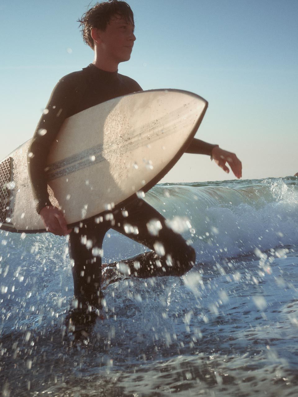 Strandszene - Ein junger Mann watet mit einen Surfbrett unter dem Arm durch das kniehohe Wasser.  