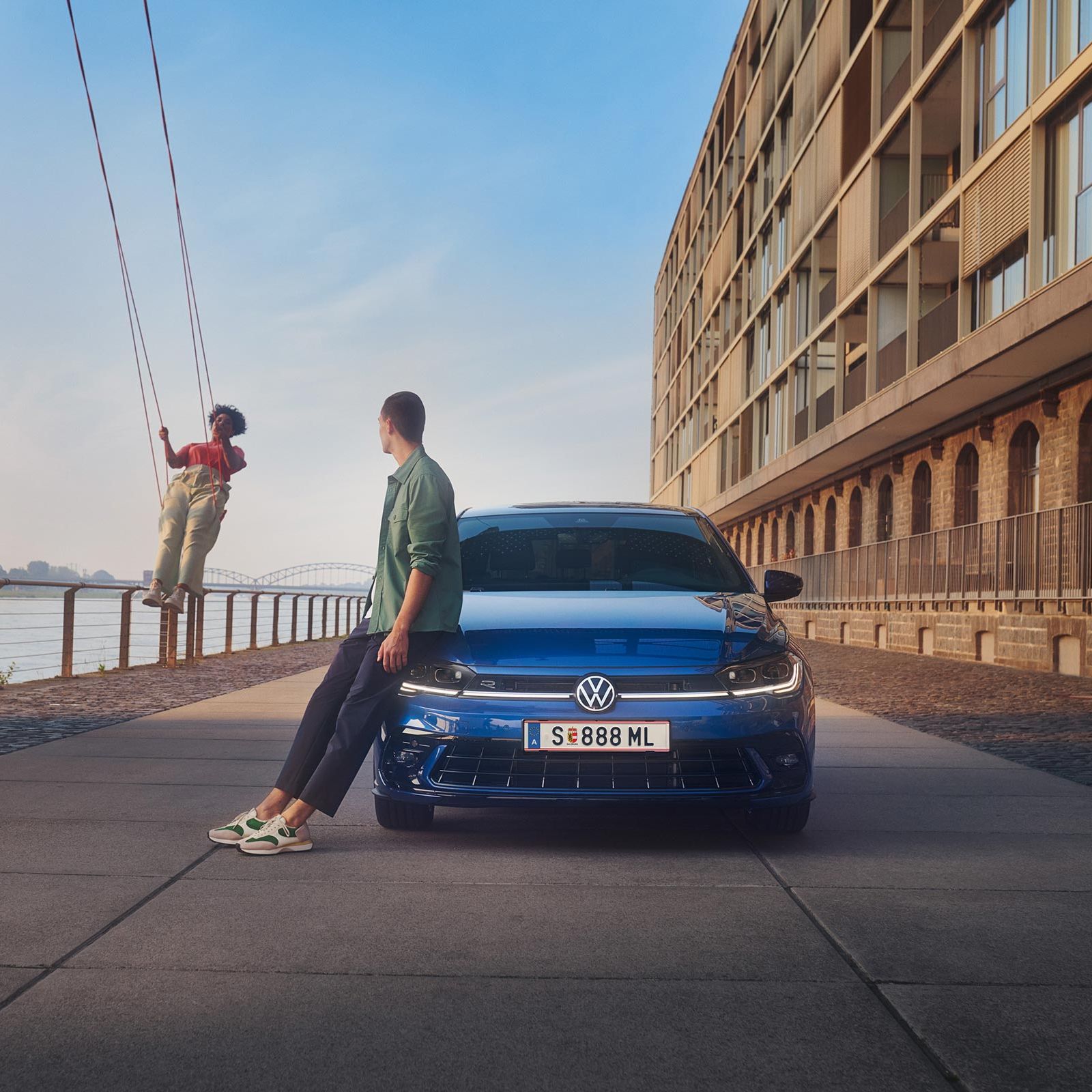 Frontansicht eines blauen VW Polo geparkt am Hafen, ein Mann lehnt an ihm, eine Frau schaukelt