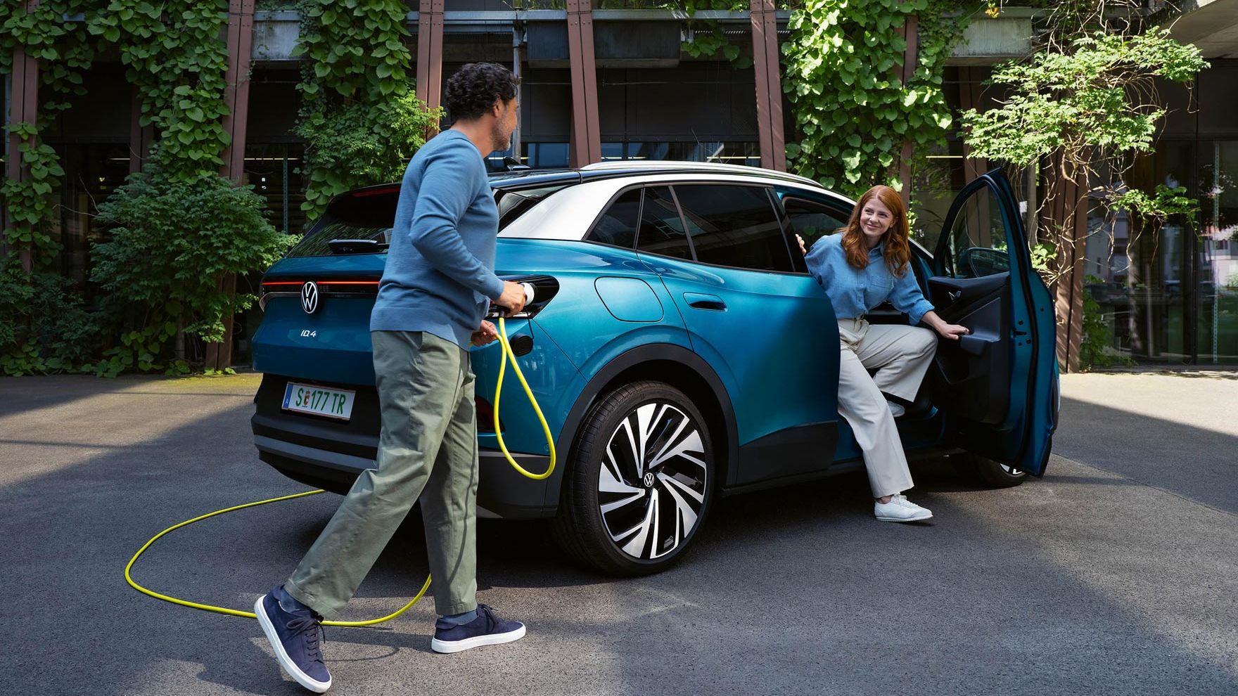 Ein Mann mit einem gelben Ladekabel für Elektroautos geht auf das Heck eines parkenden blauen VW ID.4 zu. Aus der Beifahrertür steigt eine junge Frau aus.