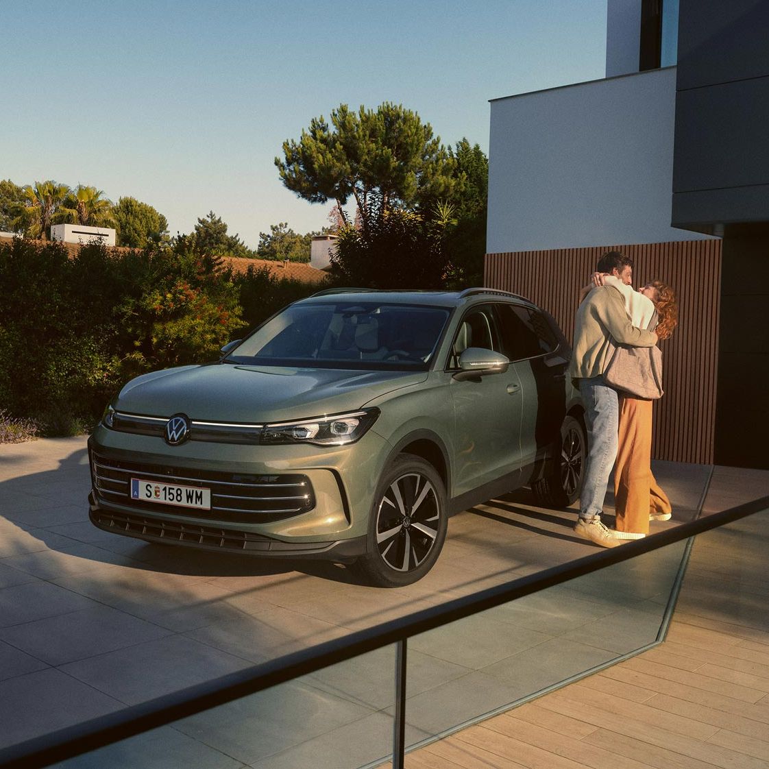 Eine Frau und ein Mann umarmen sich, neben ihnen steht ein VW Tiguan Elegance in der Farbe Cipressino-Grün Metallic