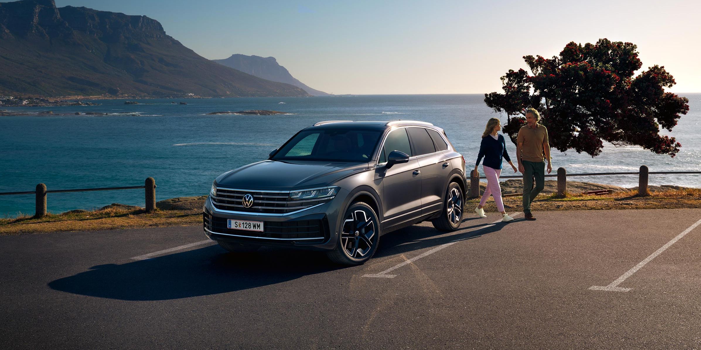 Der neue VW Touareg auf einem Parkplatz an der Küste, an ihm gehen eine Frau und ein Mann vorbei