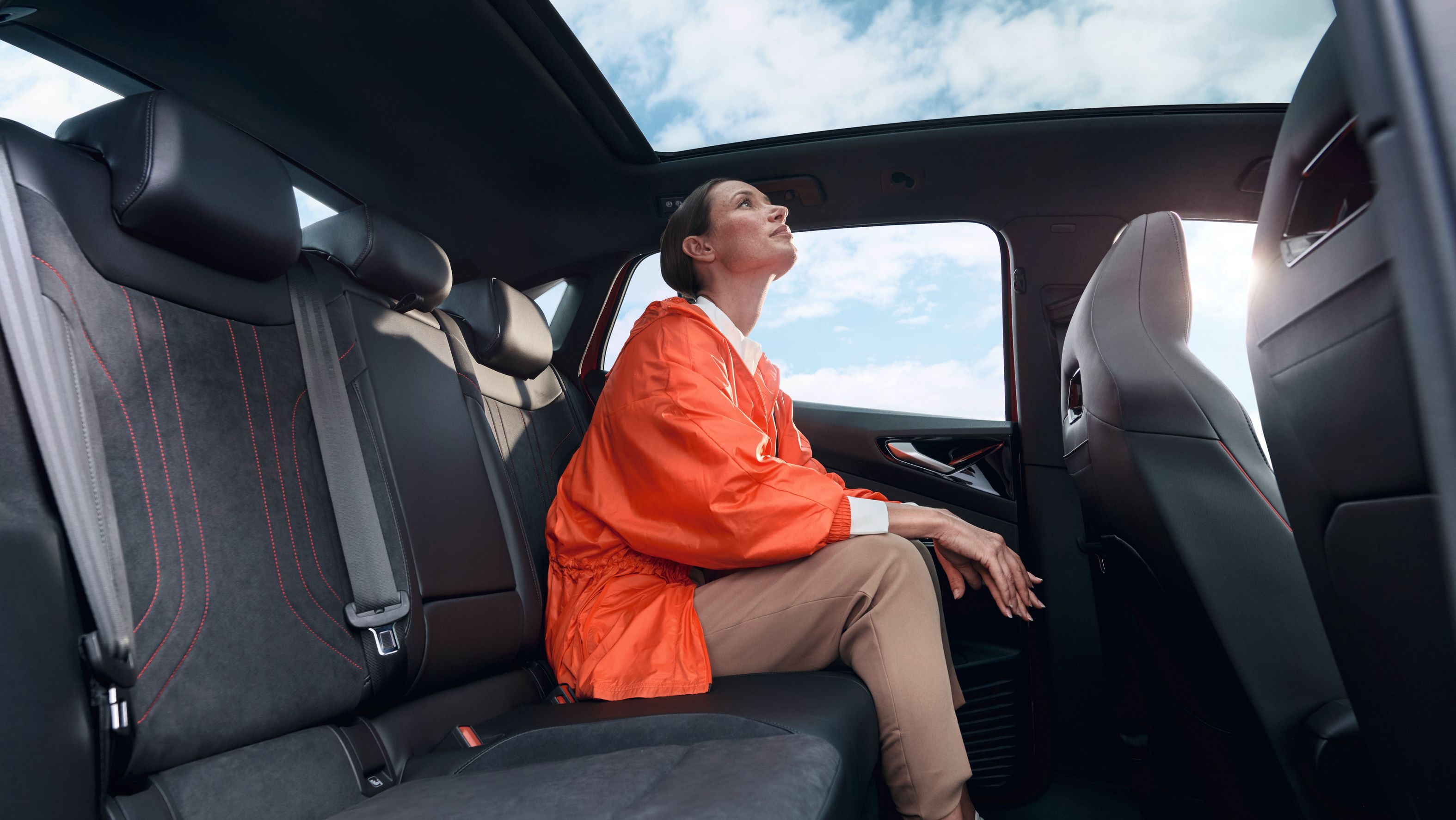 Eine junge Frau sitzt auf der Rückbank eines roten VW ID.5 GTX und schaut durch das Panoramaglasdach in den blauen Himmel.