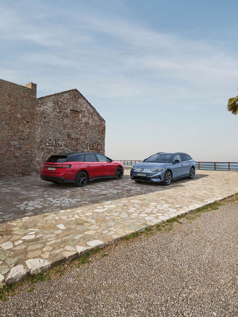 Ein VW ID.7 Tourer in Stonewashed Blue Metallic und ein VW ID.7 Tourer GTX in Kings Red Metallic geparkt an Küste neben eines Steinhaus'