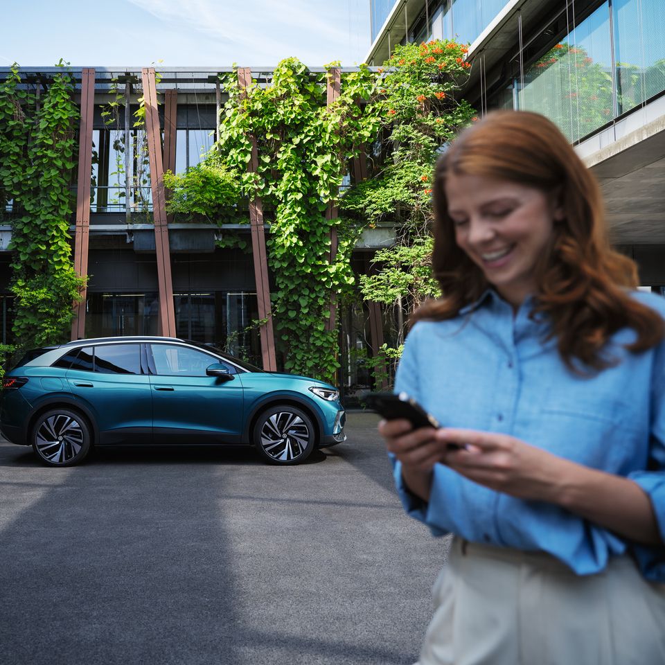 Frau lacht und blickt auf ihr Smartphone, dahinter ein blauer VW ID.4