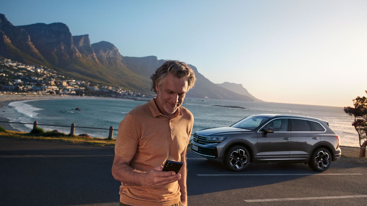 Ein Mann schaut auf sein Smartphone, dahinter parkt ein VW Touareg Elegance, im Hintergrund das Meer und die Küste.