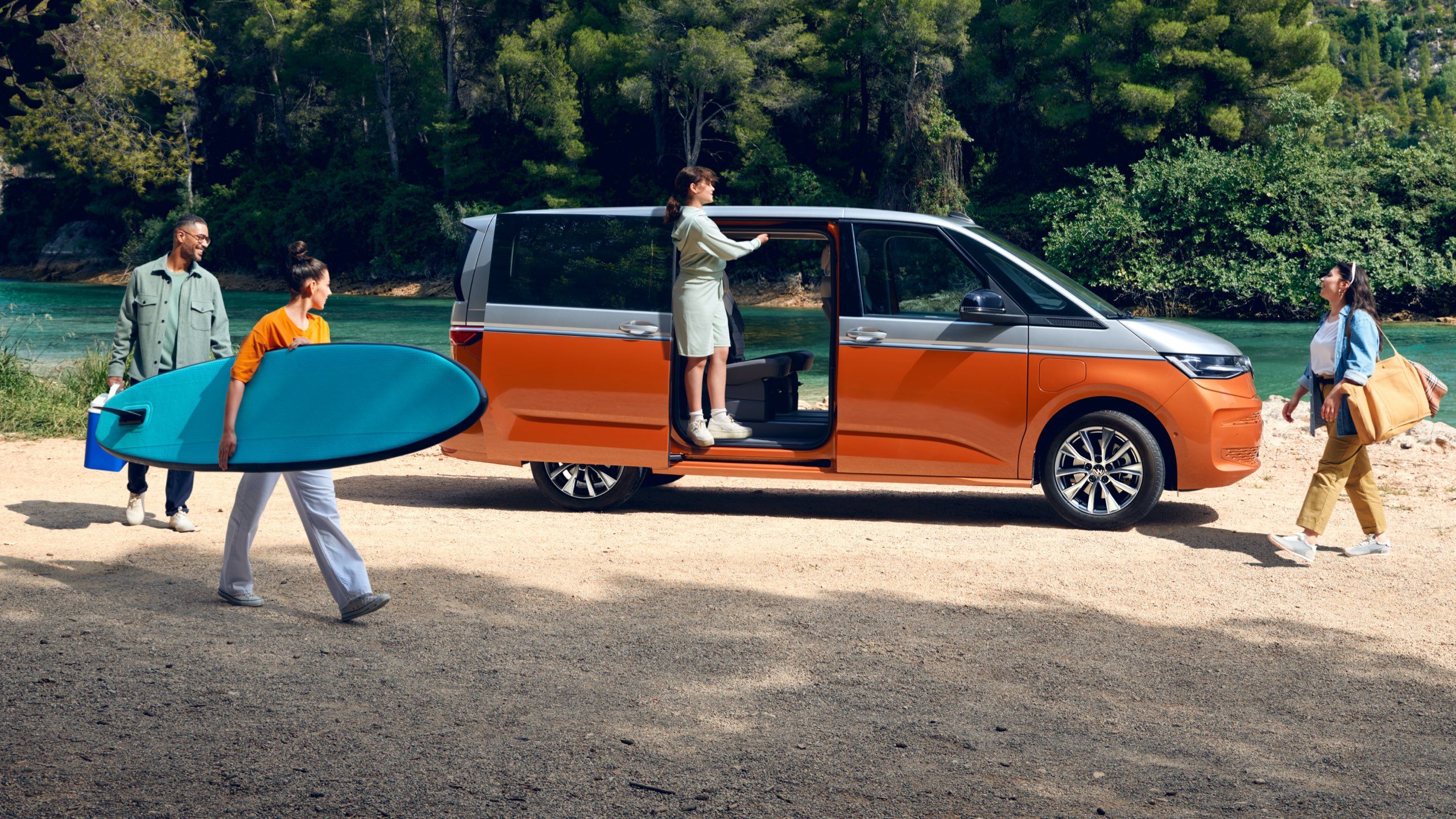 Der VW Multivan T7 Energetic steht auf einem Platz mit vier Personen vor einem Fluss. 