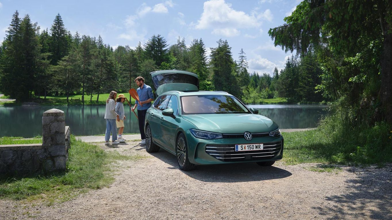 Der neue VW Passat Elegance in Maripositgrün Metallic parkt an einem Badesee mit geöffnetem Kofferraum. Ein Mann verteilt Freitzeitspielzeug an zwei Kinder.