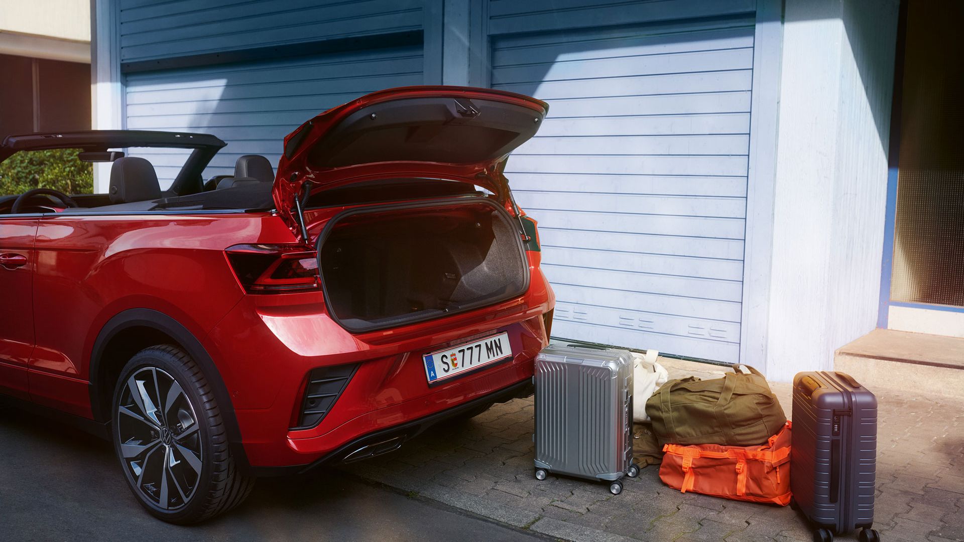 Heckansicht des VW T-Roc Cabriolet mit geöffnetem Dach und geöffnetem Kofferraum, davor stehen Koffer auf der Straße