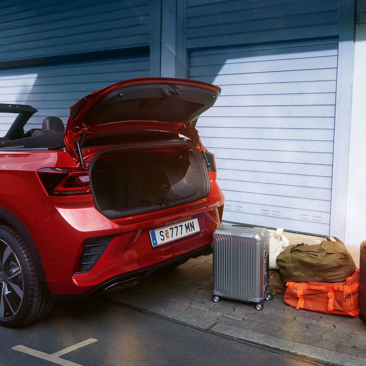 Heckansicht des VW T-Roc Cabriolet mit geöffnetem Dach und geöffnetem Kofferraum, davor stehen Koffer auf der Straße