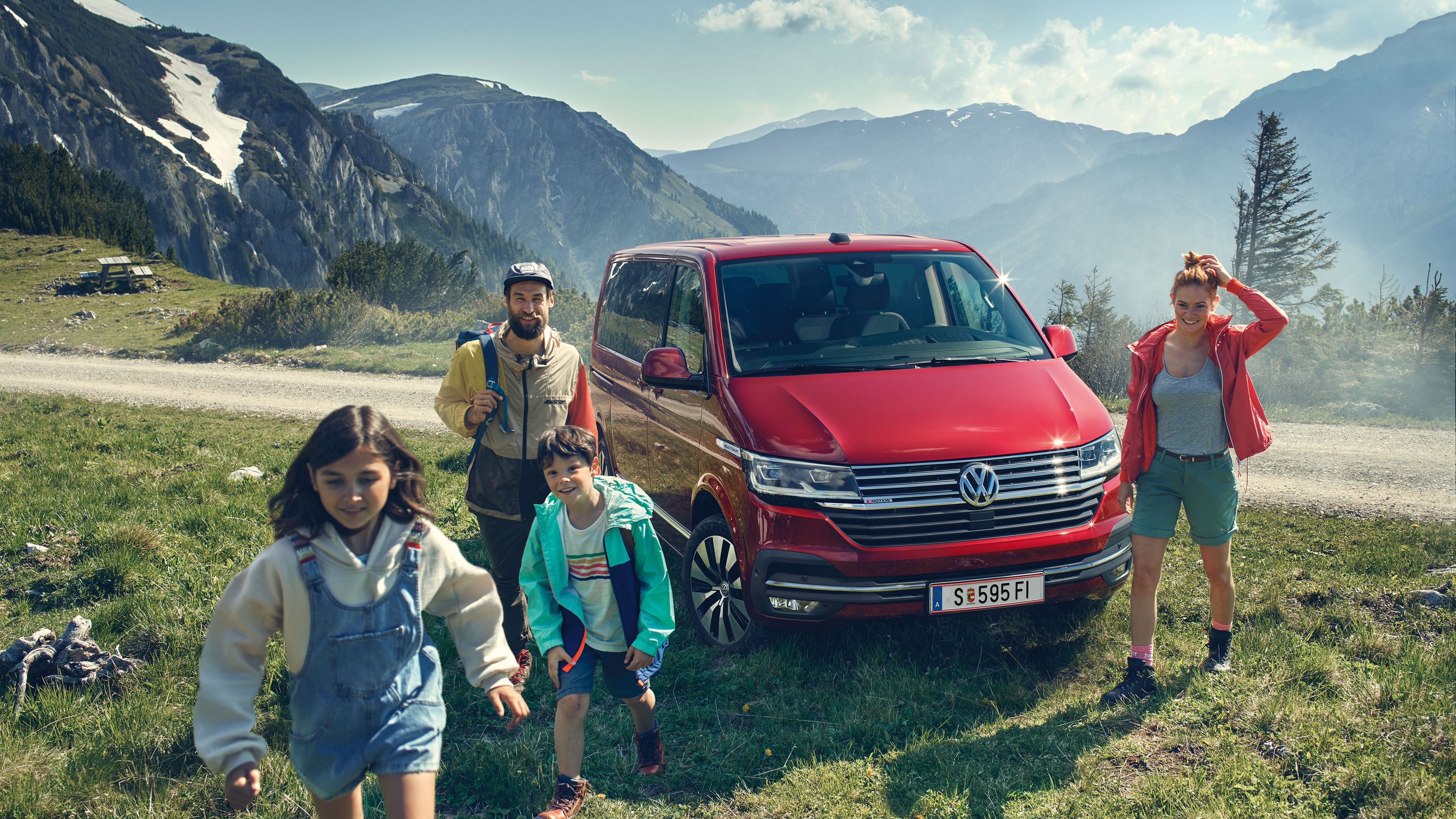 Familie geht auf Vordergrund zu. Im Hintergrund parkt ein roter VW Multivan 6.1. Dahinter eine Berglandschaft.