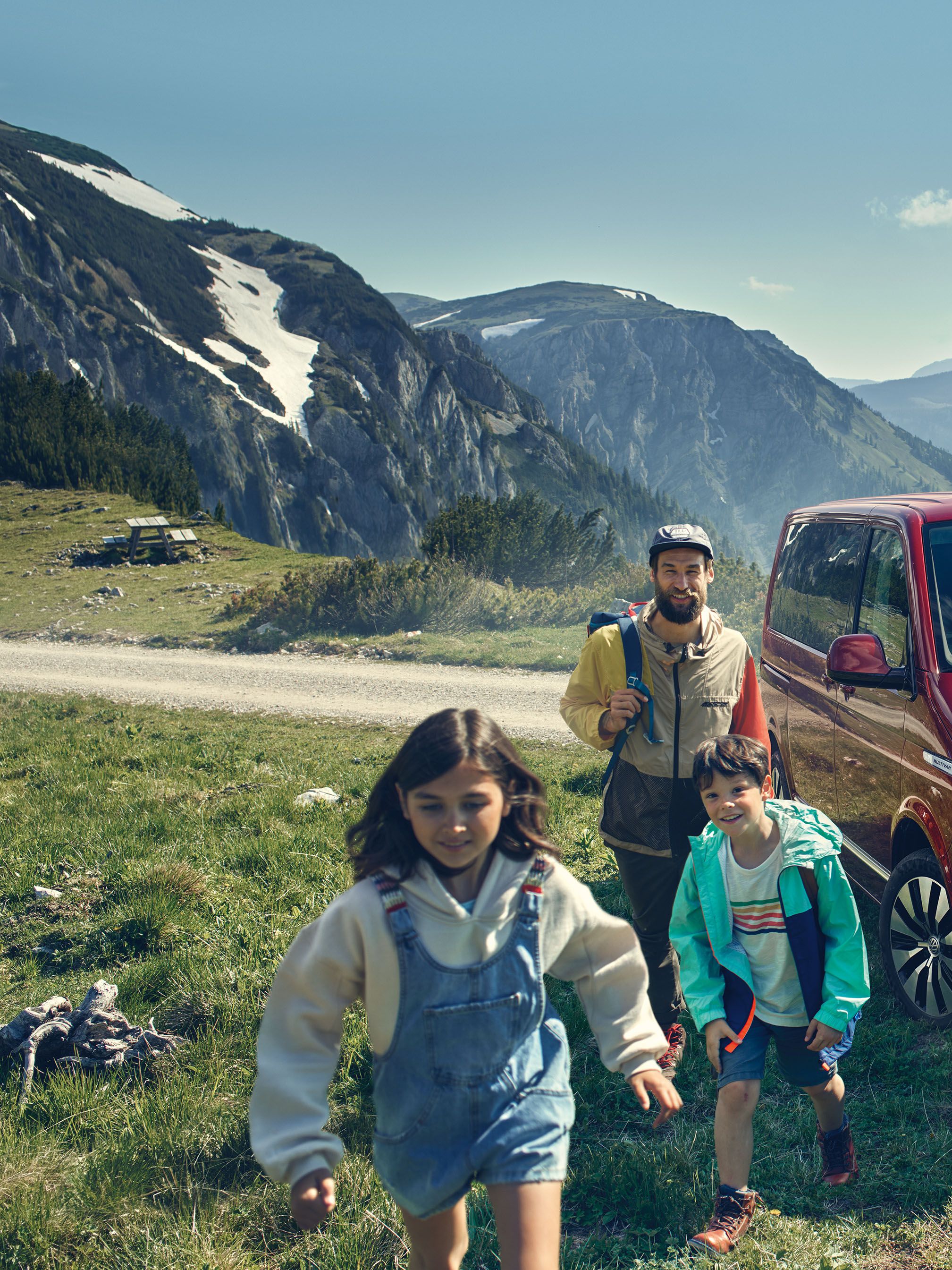 Familie geht auf Vordergrund zu. Im Hintergrund parkt ein roter VW Multivan 6.1. Dahinter eine Berglandschaft.