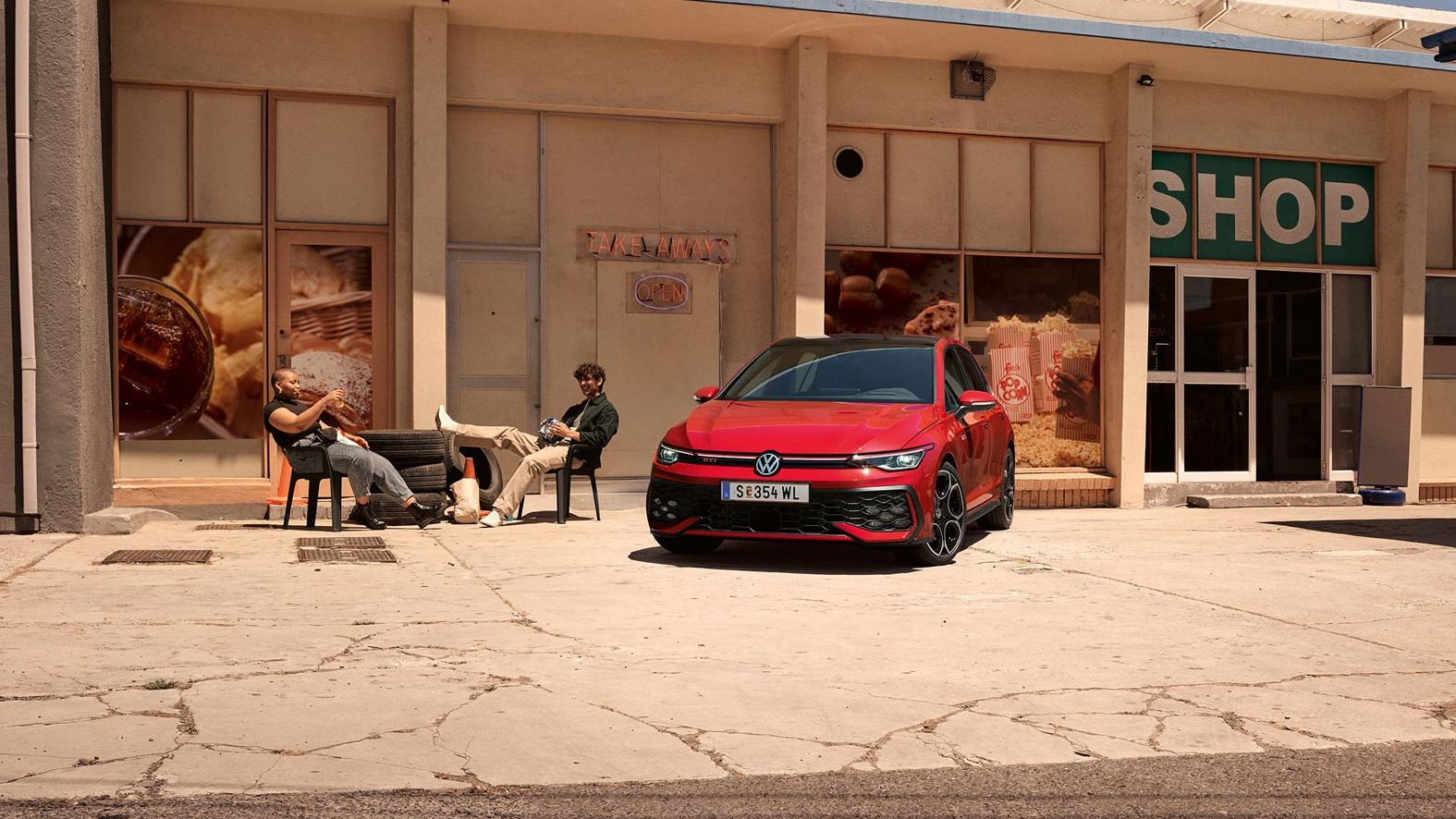 Der VW Golf GTI geparkt vor Gebäude, zwei junge Leute sitzen daneben in Stühlen