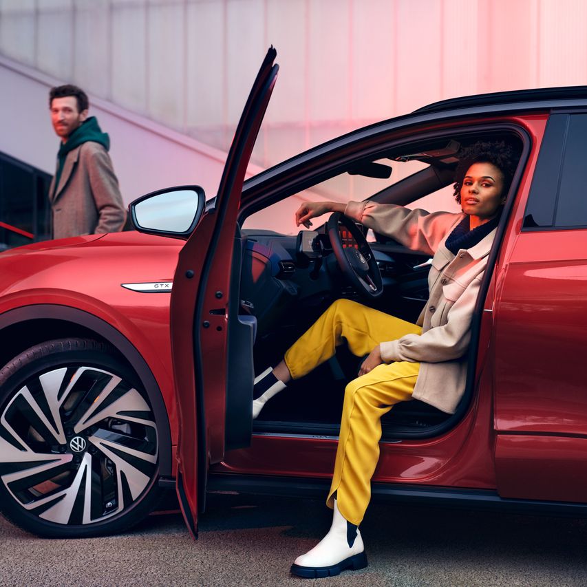 VW ID.4 GTX in Rot von der Seite sichtbar, Fahrertür geöffnet, Frau sitzt lässig auf Fahrersitz