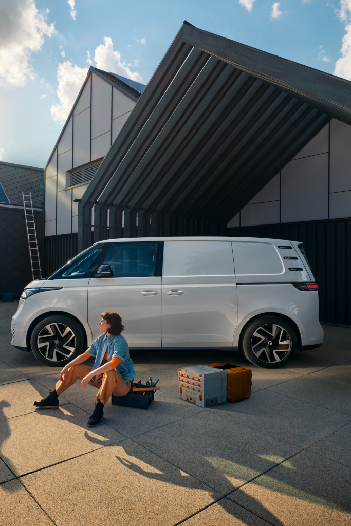 Ein weißer Volkswagen ID. Buzz Cargo vor einer Werkshalle, im Vordergrund sind ein Mann und eine Frau zu sehen