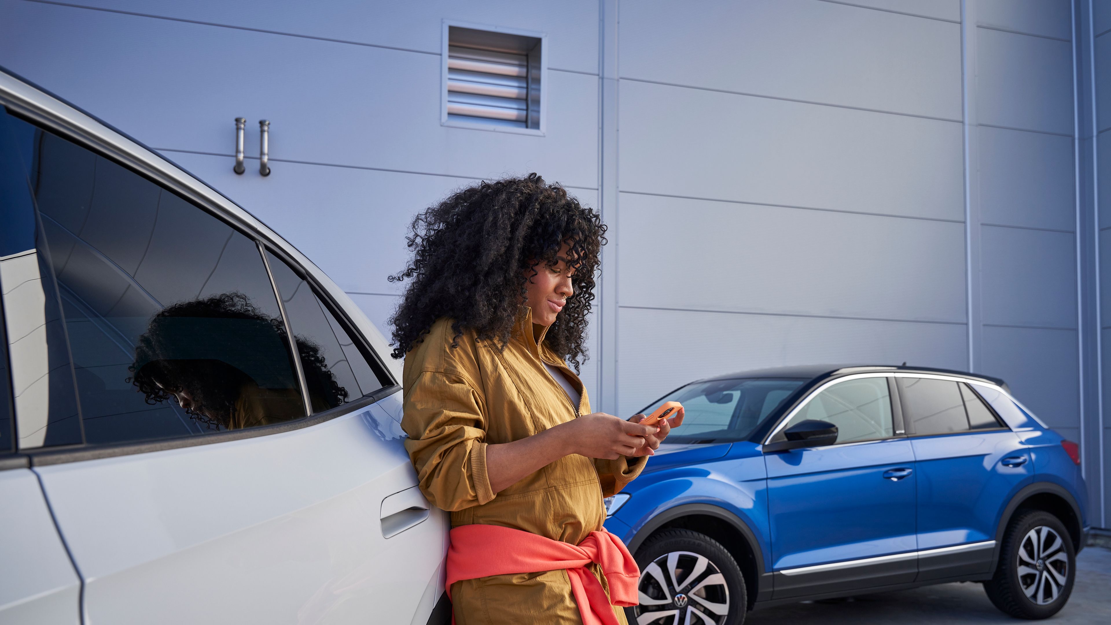 Frau lehnt an Volkswagen und blickt auf ihr Smartphone, im Hintergrund sieht man einen blauen T-Roc