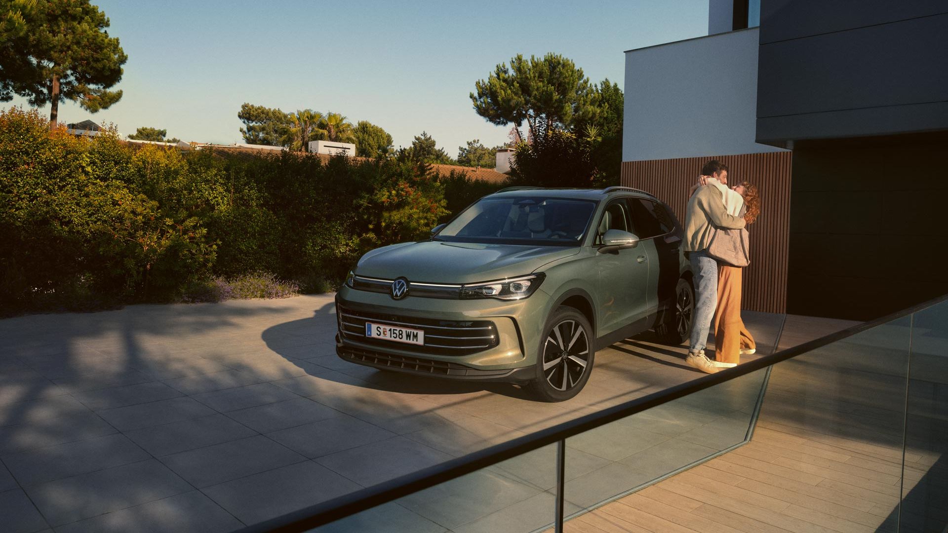 Grüner VW Tiguan parkt in Frontansicht auf der Einfahrt eines modernen Wohnhauses. Ein junges Paar steht daneben und umarmt sich.