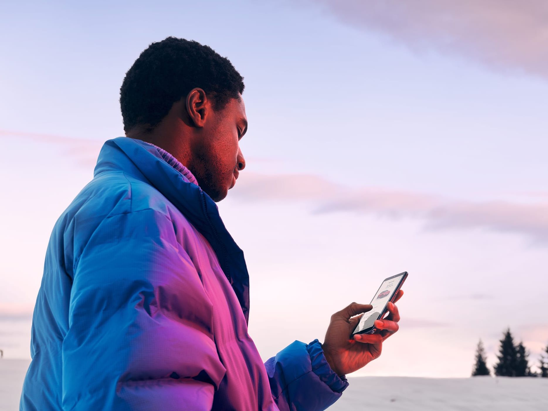 Ein Mann in Winterjacke steht in verschneiter Landschaft und schaut auf sein Smartphone.