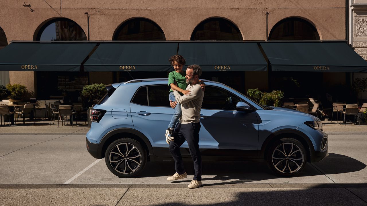 Ein Mann hält einen fröhlichen Jungen vor einem blauen VW T-Cross auf dem Arm.