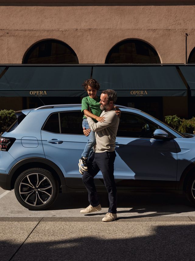 Ein Mann hält einen fröhlichen Jungen vor einem blauen VW T-Cross auf dem Arm.
